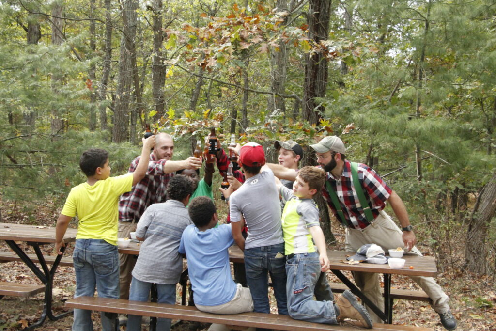 a group of boys and chiefs clinking root beer bottles at outdoor picnic